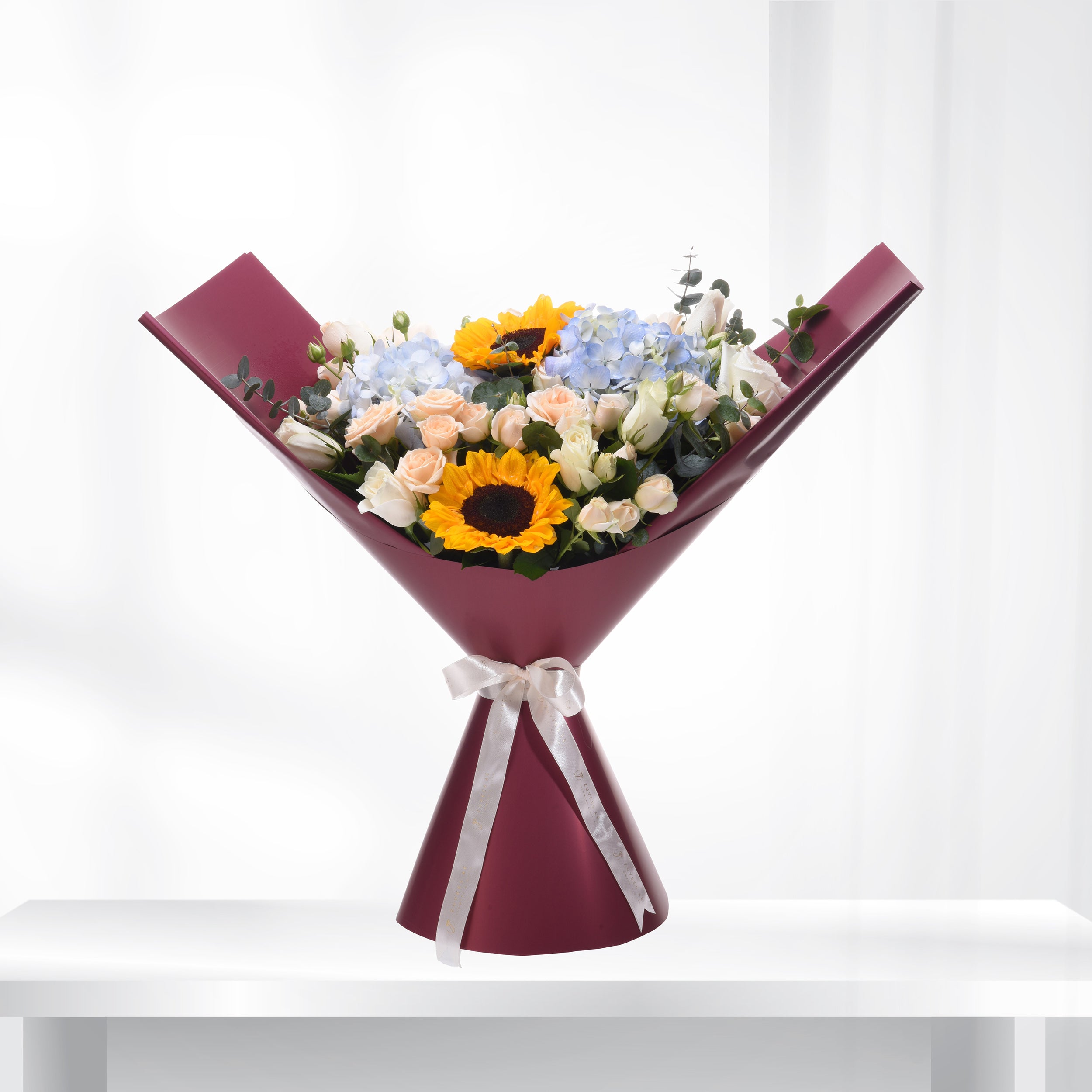 Vibrant sunflowers, roses, and hydrangeas in burgundy wrap with silver bow.