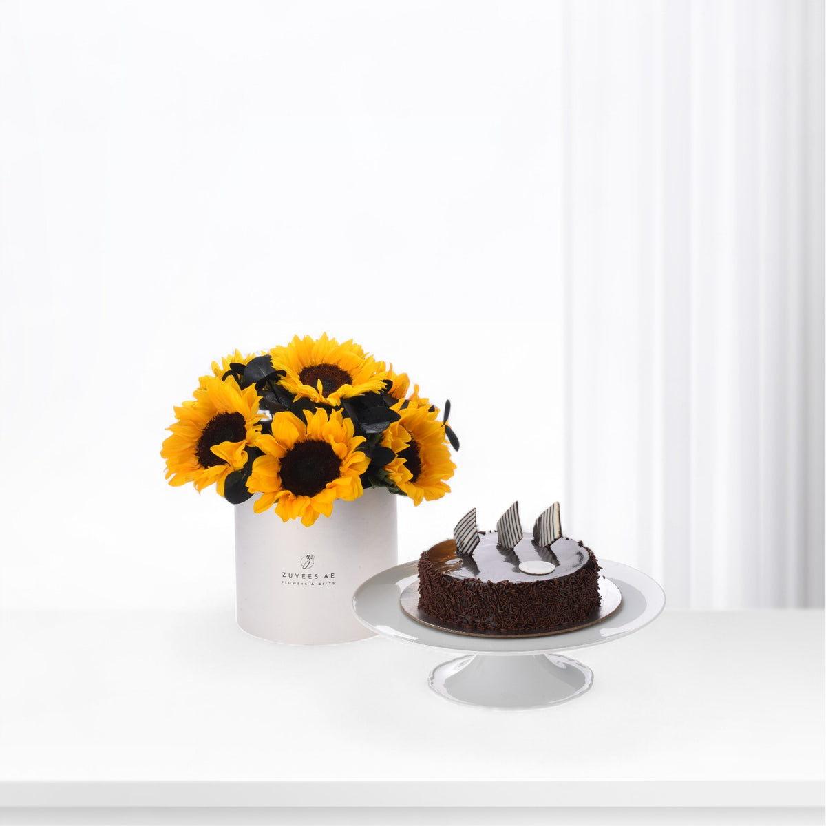 Vibrant sunflowers in white vase beside chocolate cake on stand.