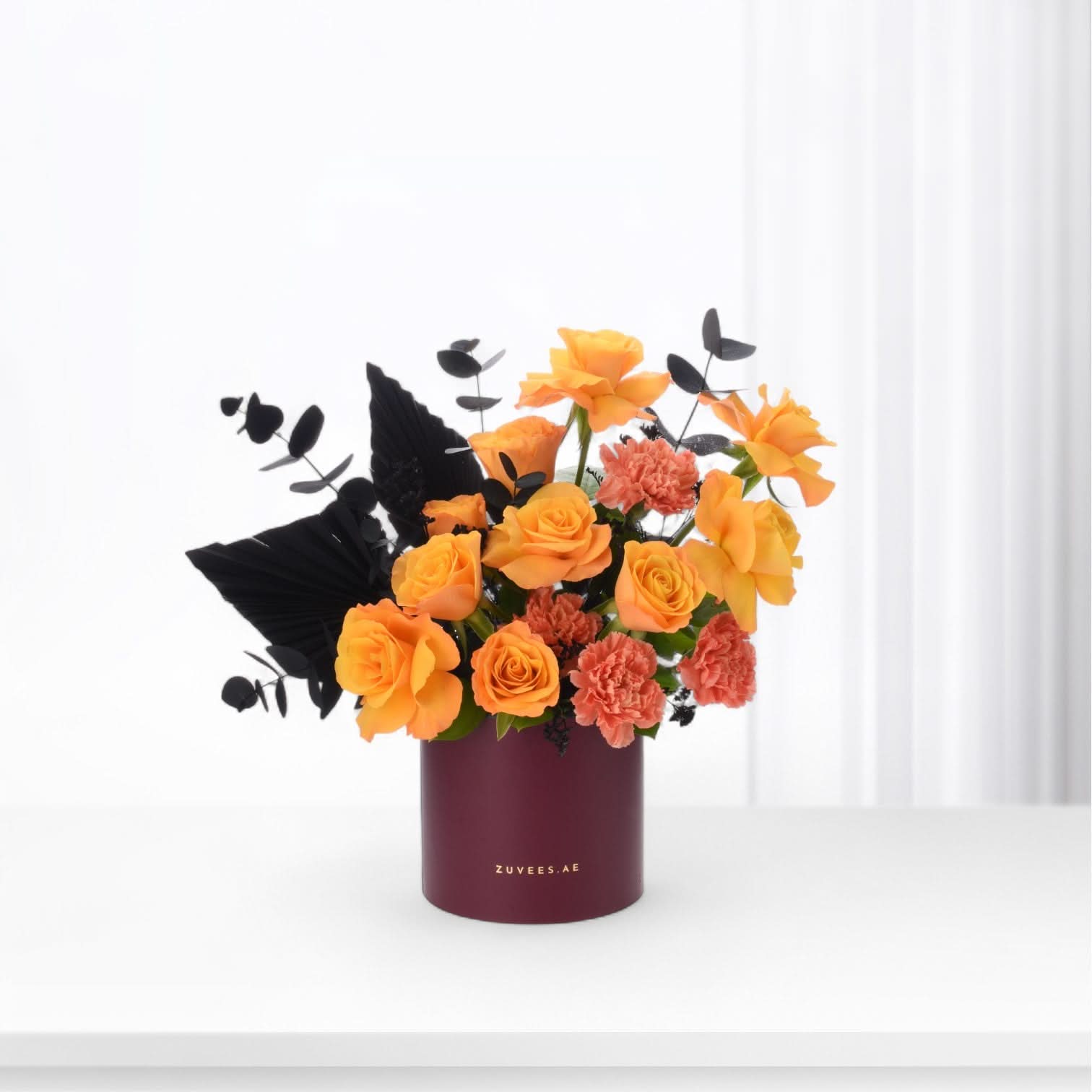 Vibrant orange roses and carnations contrast elegantly with black foliage in a burgundy cylinder vase.