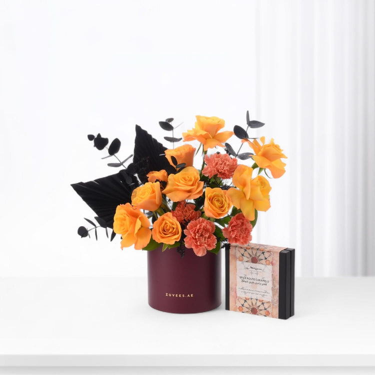 Vibrant orange roses and carnations in burgundy vase beside caramel gift box.