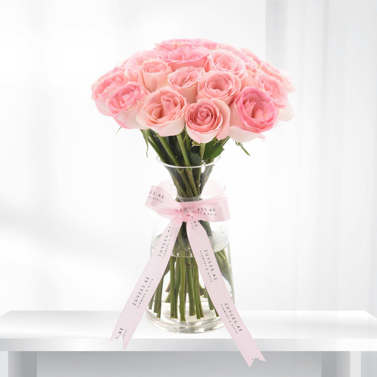 Delicate pink roses in clear vase, tied with branded ribbon, soft white background.