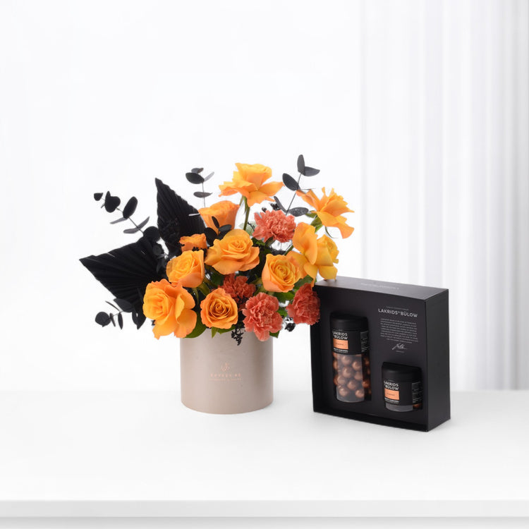 Vibrant orange roses and black foliage in beige vase beside sleek black chocolate gift box.