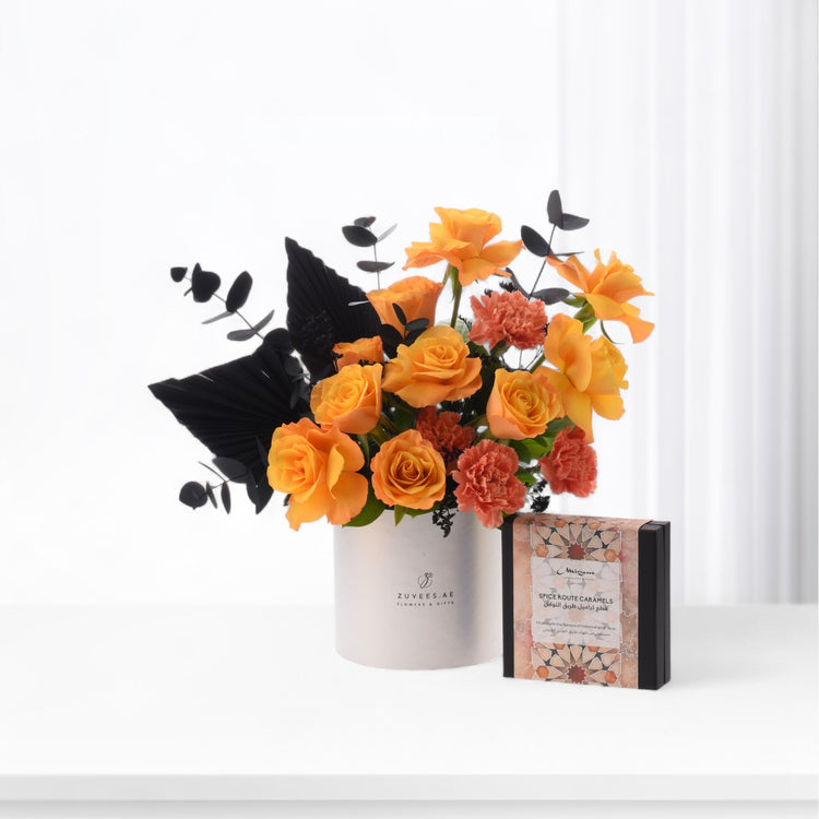 Vibrant orange roses and coral carnations contrast bold black foliage in sleek white vase beside spice caramel gift box.