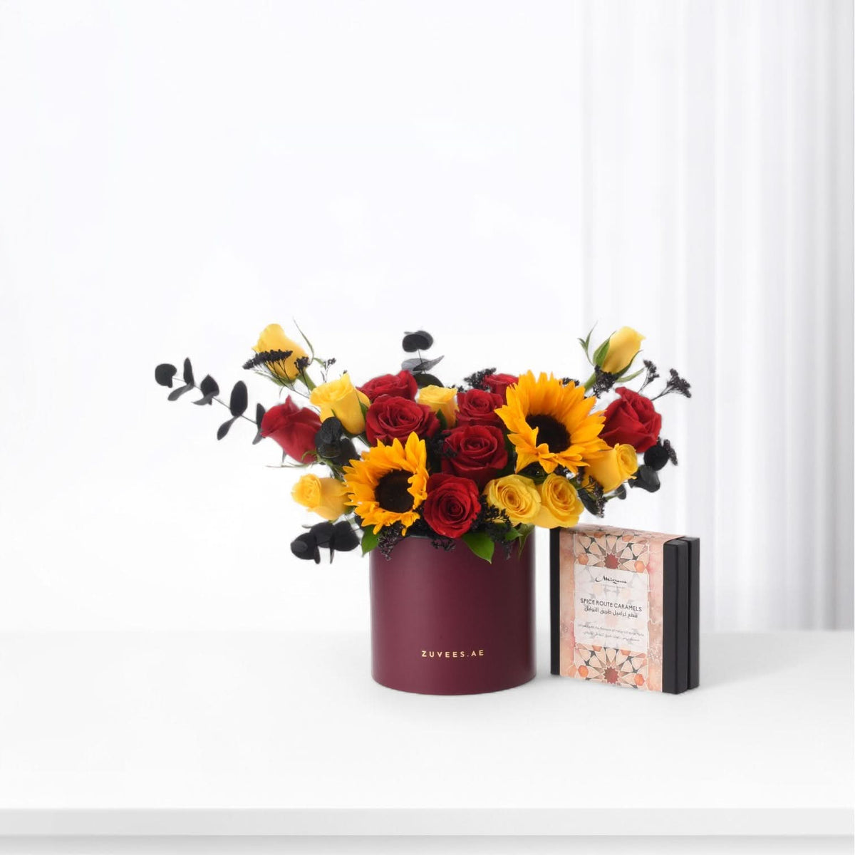 Vibrant red and yellow roses with sunflowers in burgundy vase beside caramel gift box.
