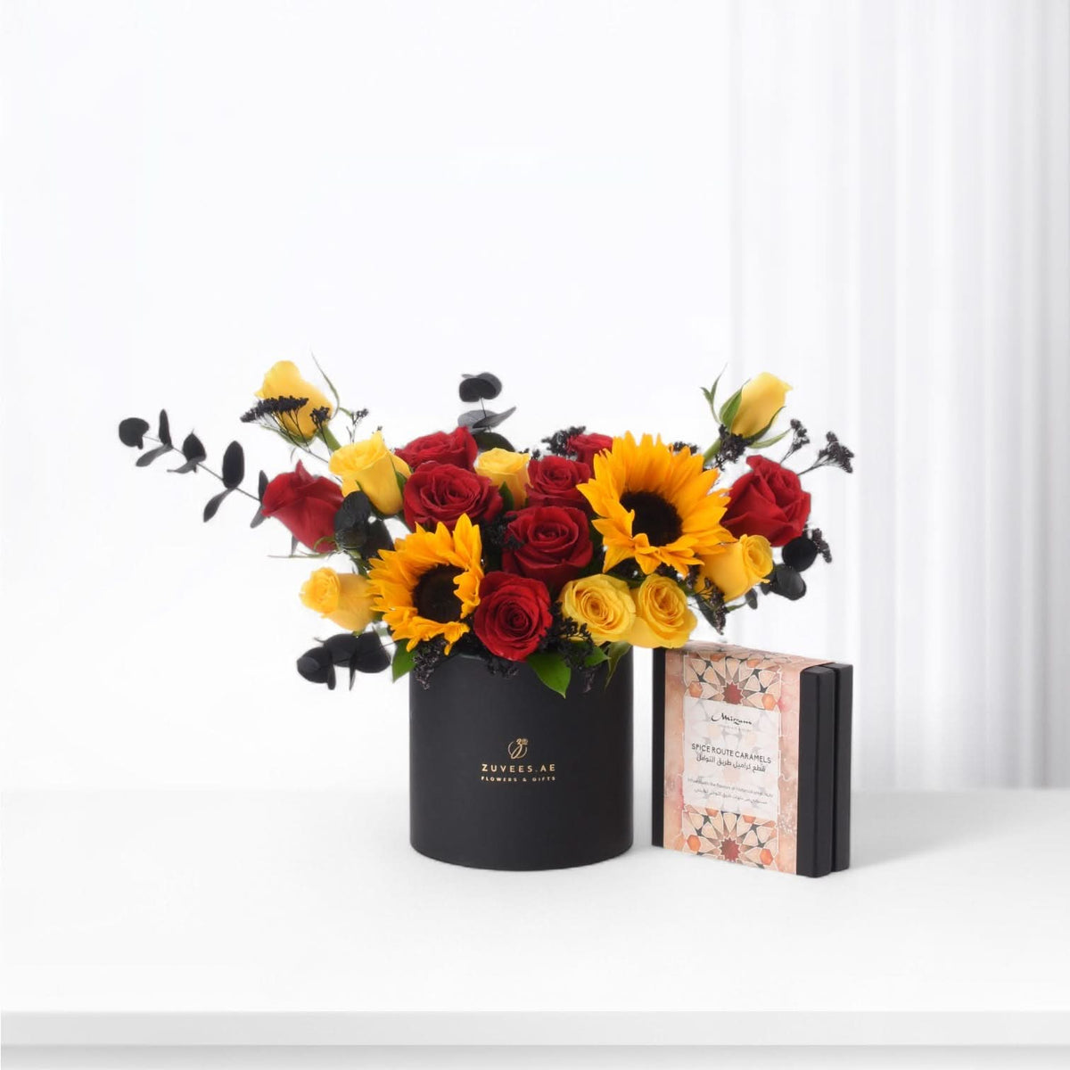 Vibrant red roses, yellow sunflowers, and black eucalyptus in sleek black vase beside caramel gift box.