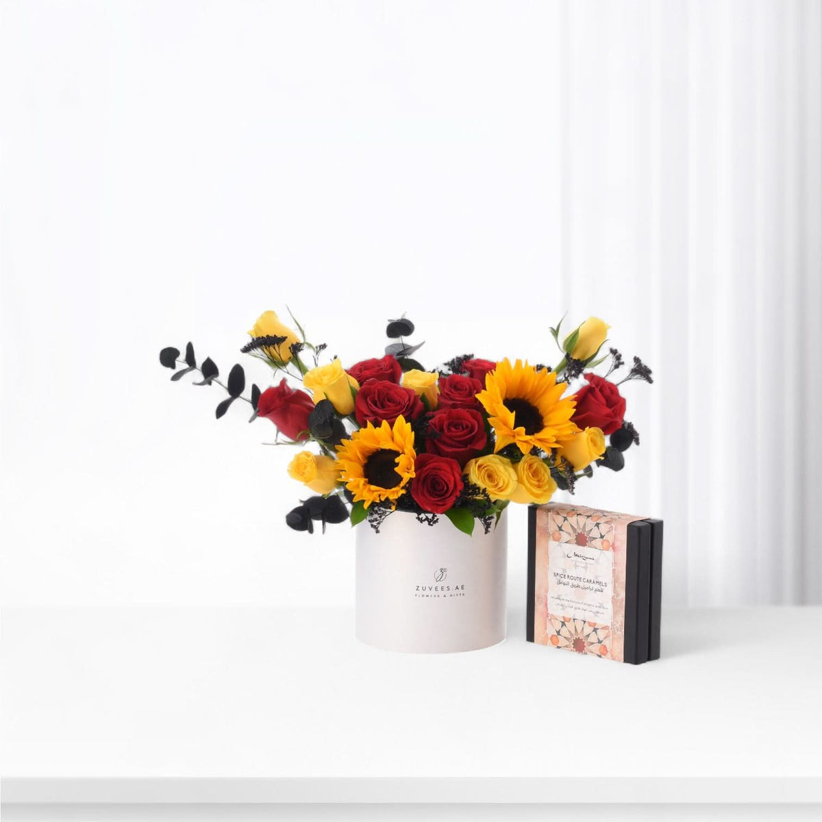 Vibrant red roses, yellow sunflowers, and black eucalyptus in sleek white box beside caramel gift.