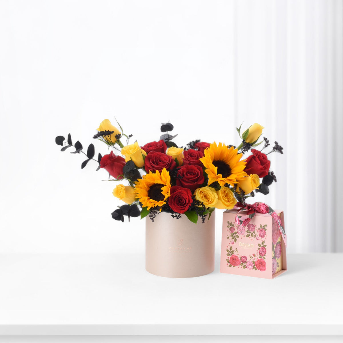 Vibrant red and yellow roses with sunflowers in rose-gold vase beside floral gift box.