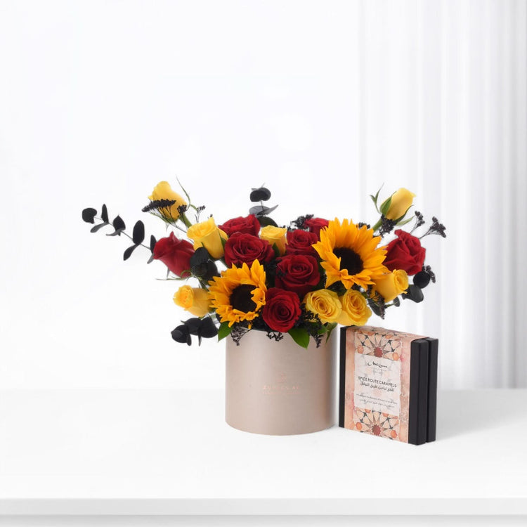 Vibrant red and yellow roses with sunflowers in beige vase, beside caramel gift box.
