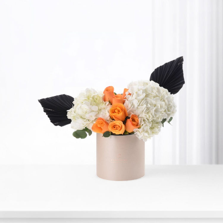 Vibrant orange roses, white hydrangeas, black leaves in chic beige cylinder vase.