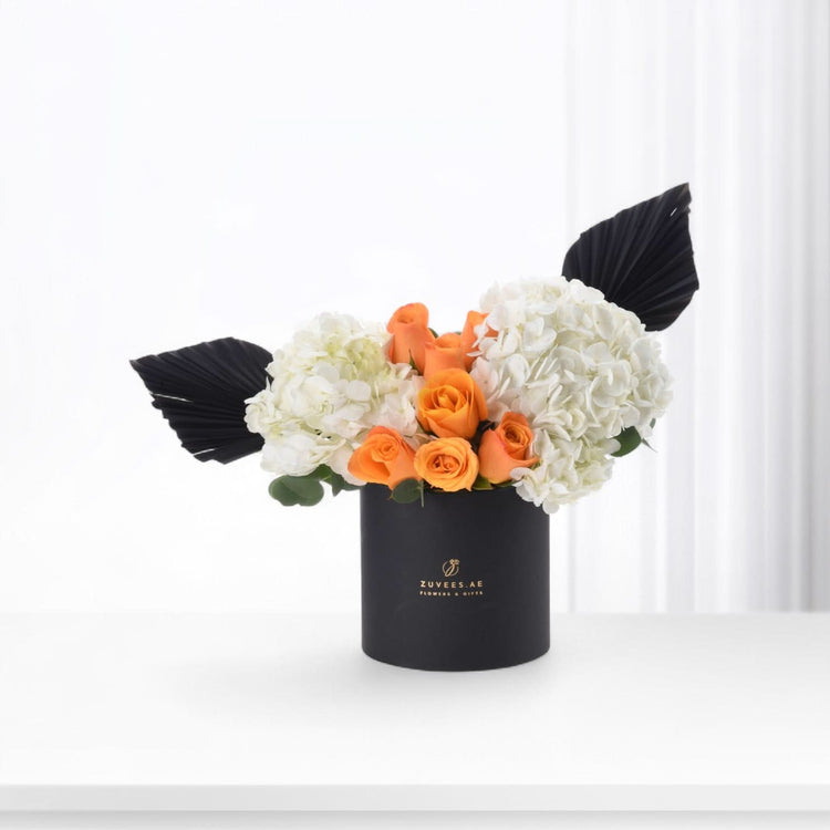 Black cylindrical vase holds vibrant orange roses and white hydrangeas, accented with dramatic black fan leaves.