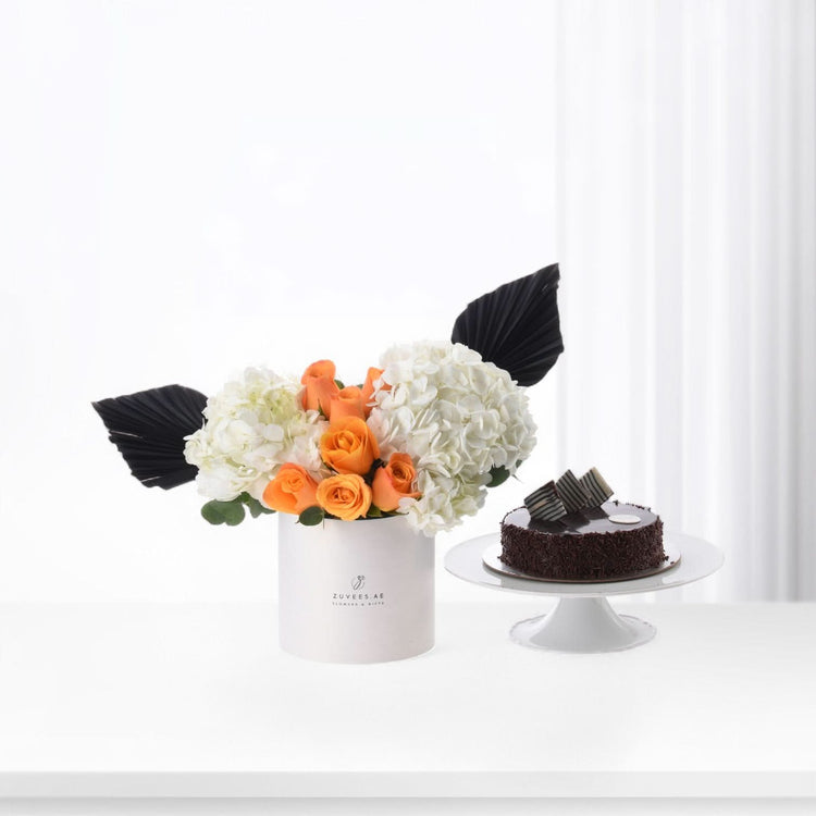 White box with orange roses and white hydrangeas, accented by black fan leaves, beside chocolate cake.