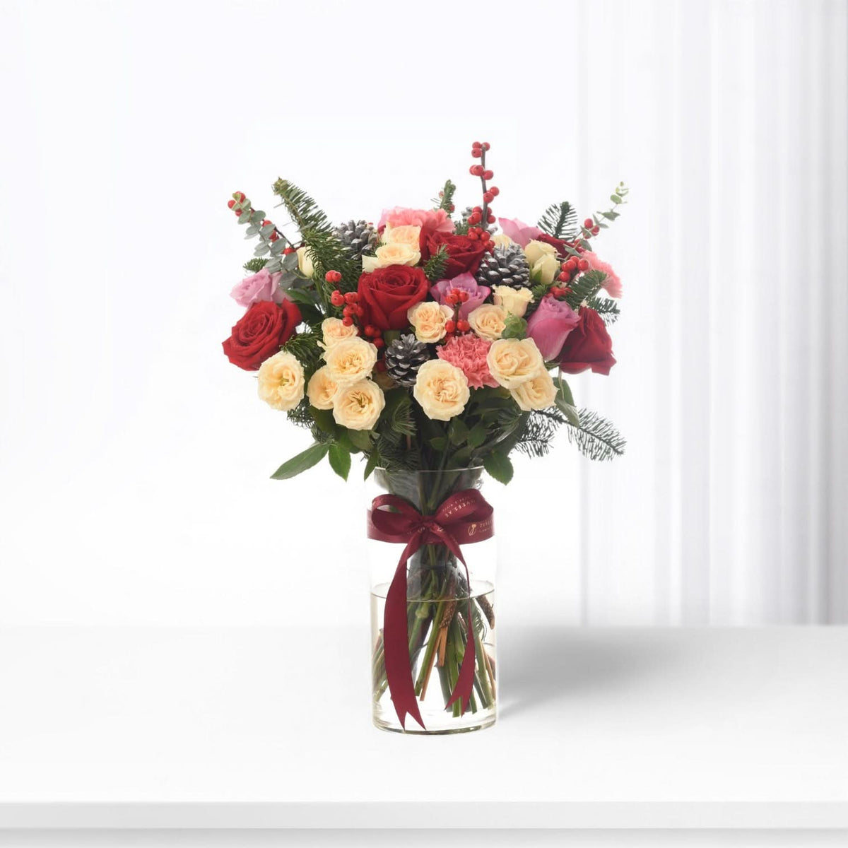 Vibrant red, pink, and cream roses with pinecones and berries in clear glass vase.