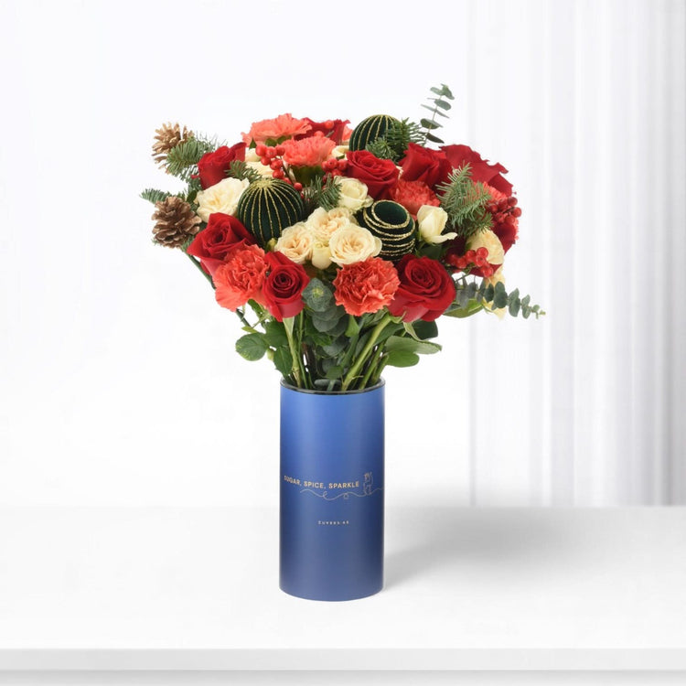 Vibrant red, cream roses, berries, pine, and ornaments in sleek blue vase.