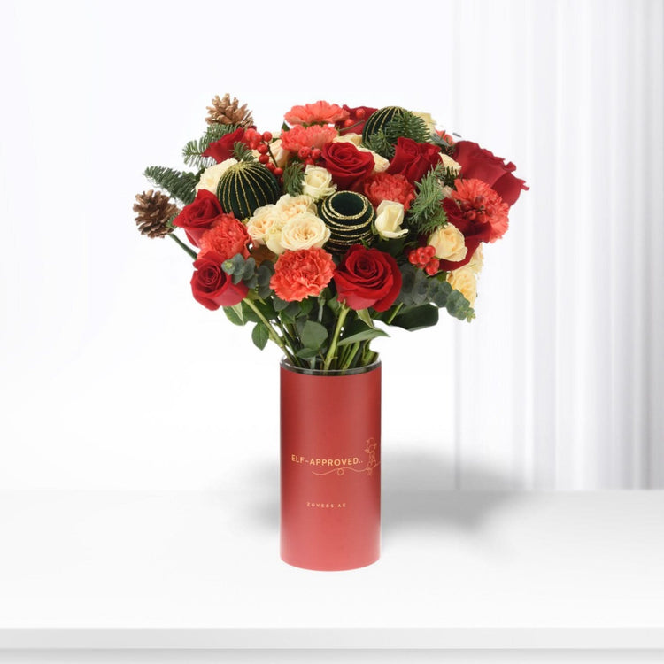 Vibrant red and cream roses, pinecones, and greenery in festive red cylinder vase.