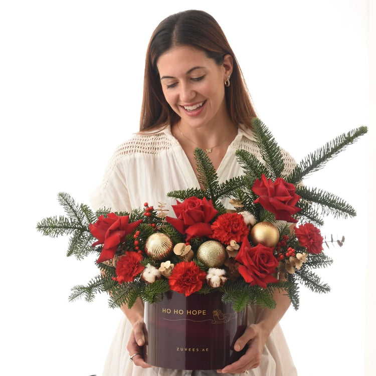 Woman holds festive red rose and pine arrangement in maroon "Ho Ho Hope" box.