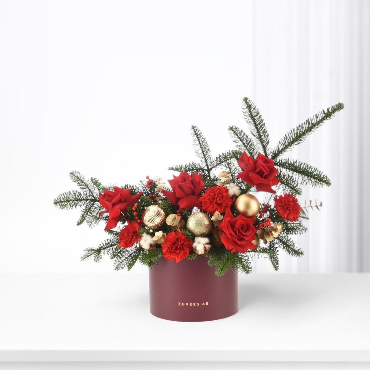 Vibrant red roses and carnations nestled in lush greenery, accented with gold ornaments, presented in a burgundy cylindrical vase.