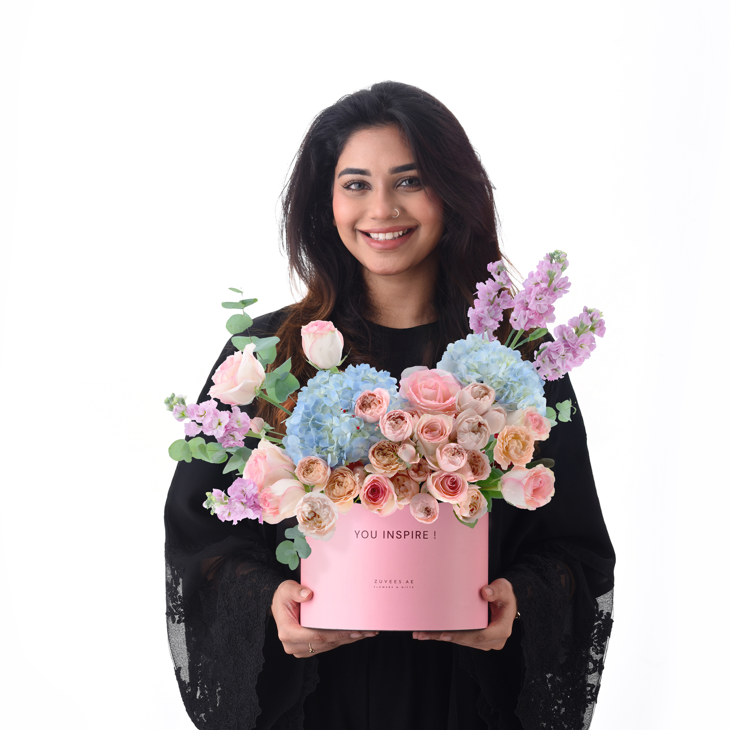 Pink box holds pastel roses, hydrangeas, and stock; woman smiles warmly.