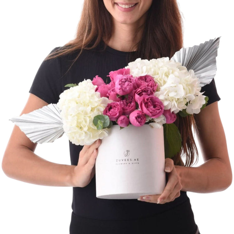 White cylindrical vase holds vibrant pink peonies and white hydrangeas, accented with silver fan embellishments.