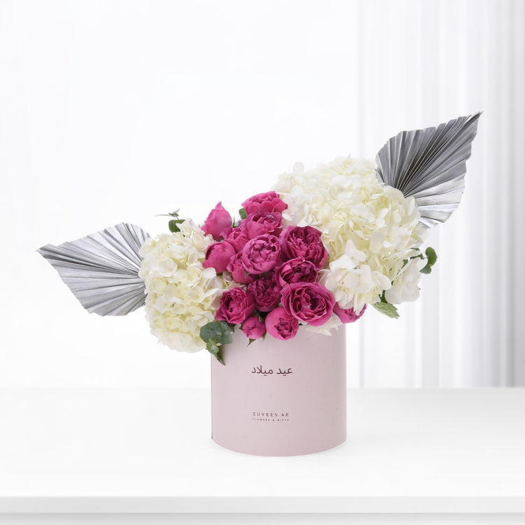 Pink box holds vibrant magenta roses and creamy hydrangeas, accented by silver fan leaves.