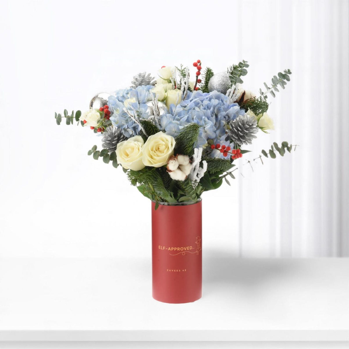 Red vase holds festive blue hydrangeas, white roses, pine, and silver ornaments.