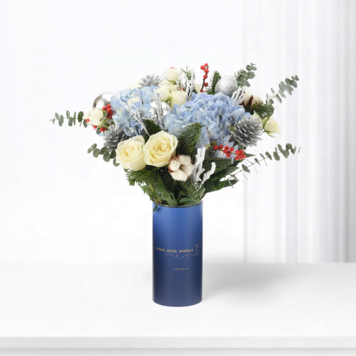 Blue vase holds snowy white roses, blue hydrangeas, silver pinecones, and red berries.