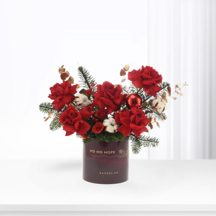 Vibrant red roses, pine, cotton, and ornament in festive deep red vase.