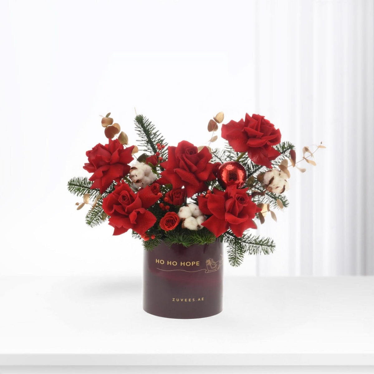 Vibrant red roses, pine, cotton, and ornament in festive deep red vase.