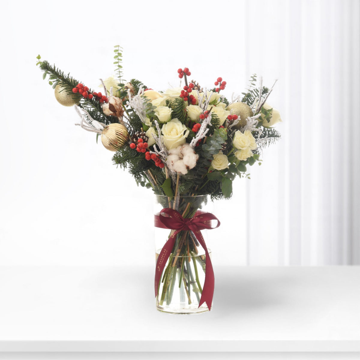 Elegant Christmas bouquet with white roses, red berries, gold ornaments, and fir in clear vase with burgundy ribbon.