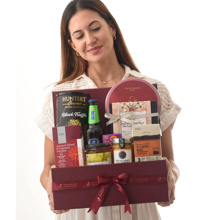 Woman holds burgundy gift box filled with gourmet treats and branded ribbon.