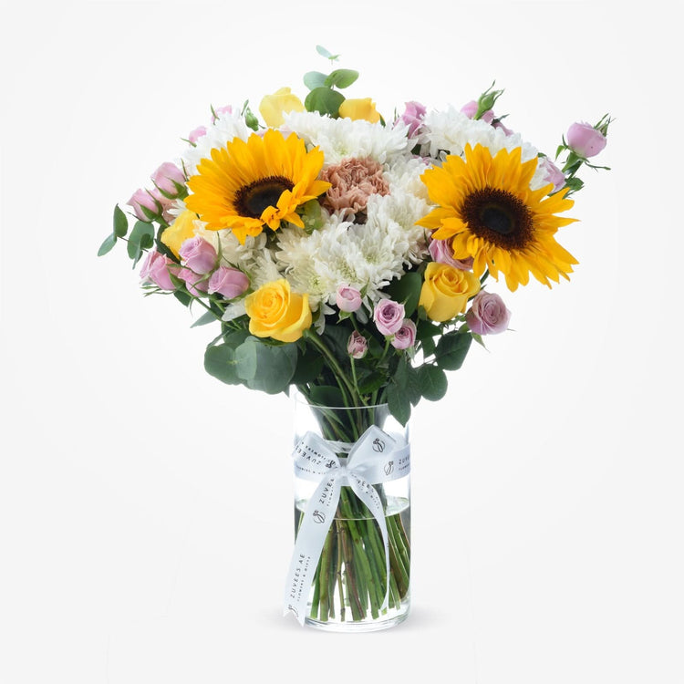 Vibrant sunflowers, roses, and chrysanthemums in clear vase with branded bow.