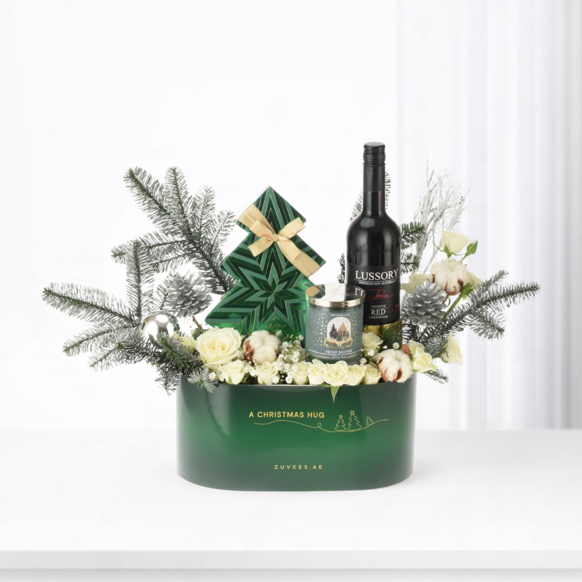 Green gift basket with white roses, pine, candle, and wine bottle.