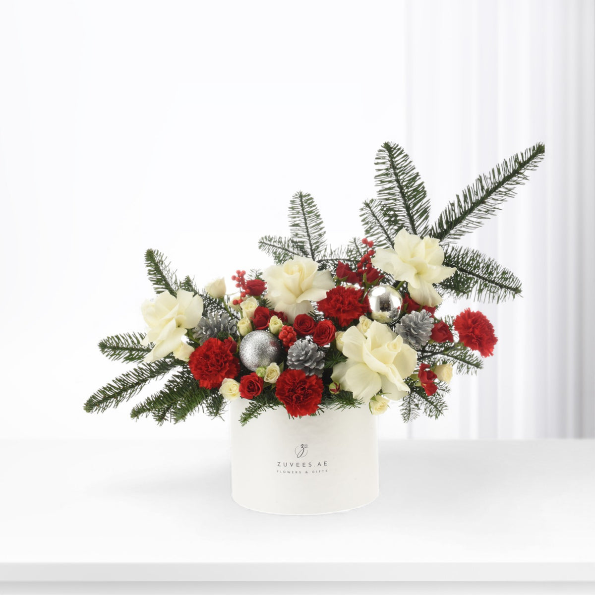White cylindrical vase holds festive red and cream blooms with pine, silver ornaments, and berries.