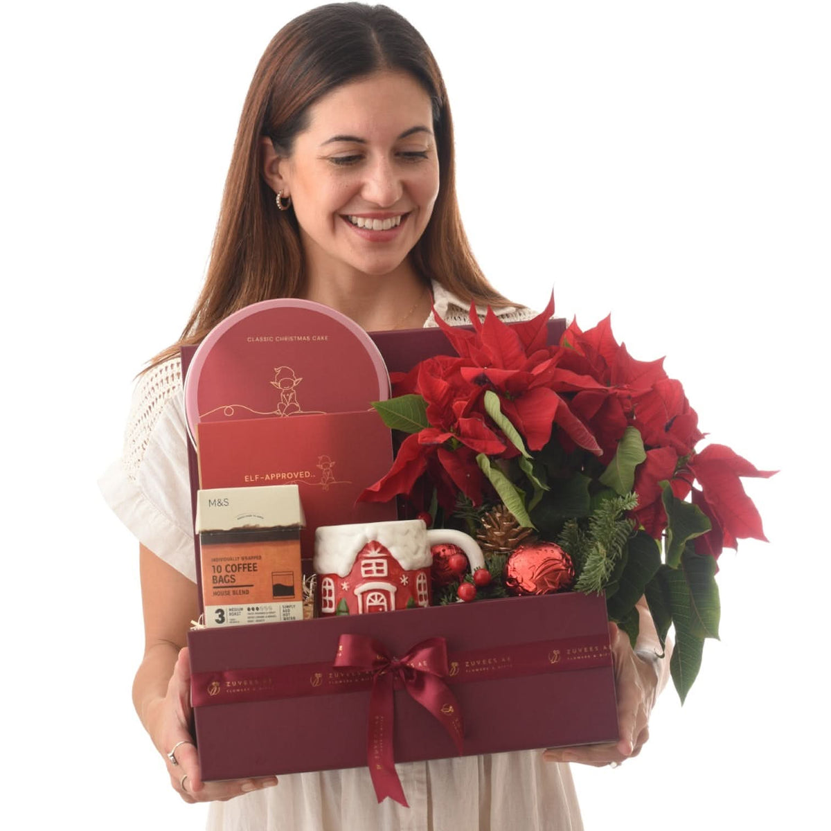 Woman smiles holding festive red gift box with poinsettias, cake, coffee, and mug.