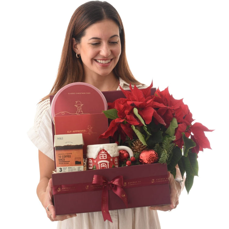 Woman smiles holding festive red gift box with poinsettias, cake, coffee, and mug.