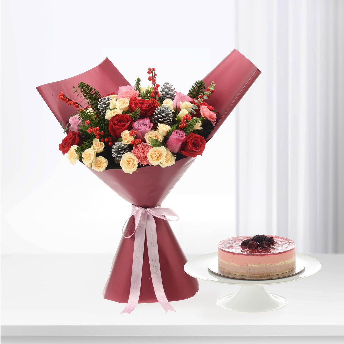 Vibrant red, pink, and cream roses wrapped in burgundy paper beside berry-topped pink cake.