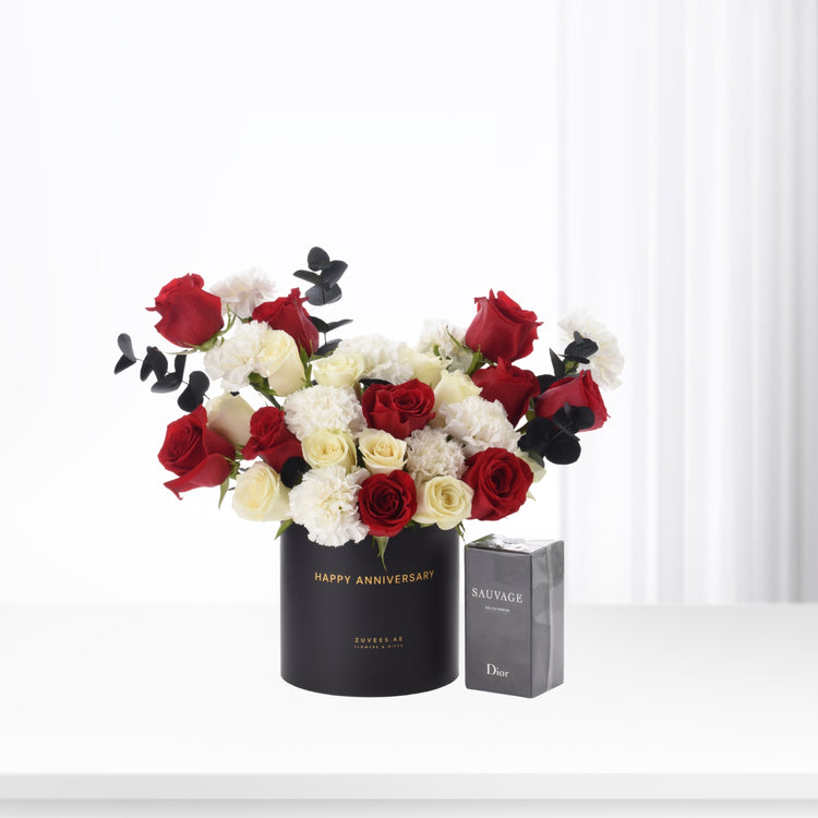 Elegant red, white roses and carnations in black "Happy Anniversary" vase beside Dior Sauvage perfume.