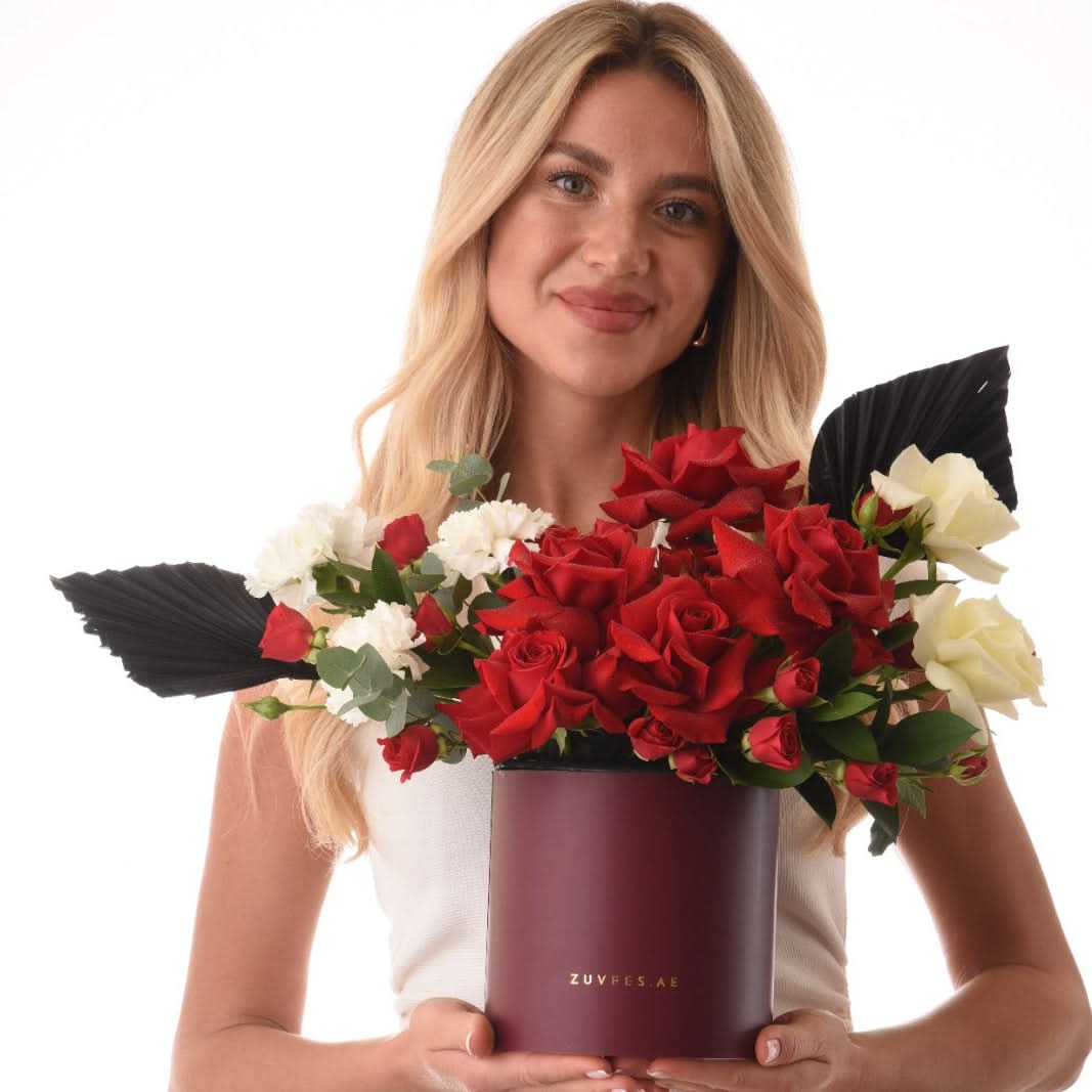 Blonde woman holds burgundy Zuvfes box with red, white roses, black leaves, greenery against white.