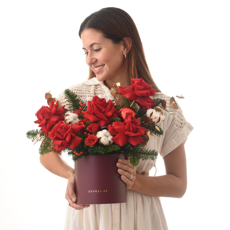 Woman holds festive red rose bouquet with pine, cotton, berries in burgundy Zuvees box.