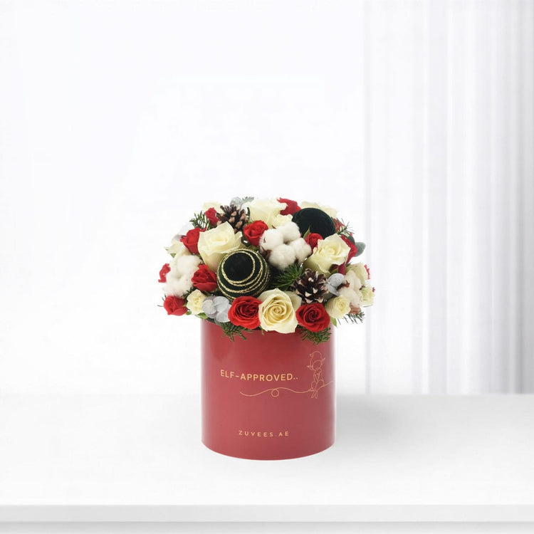 Red cylindrical box holds festive red and white roses, cotton, pinecones, and gold-accented ornaments.