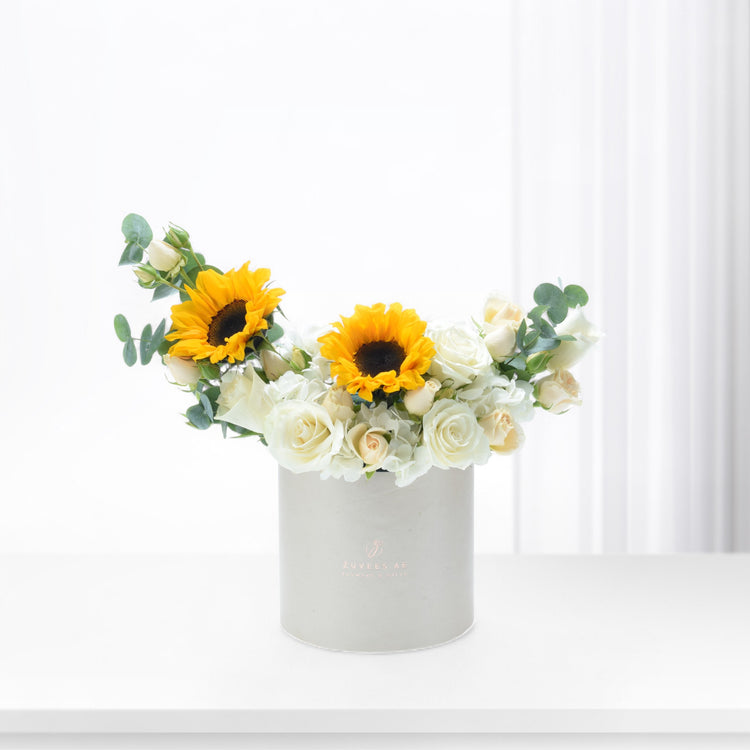 White cylindrical box holds vibrant sunflowers and creamy roses against bright background.