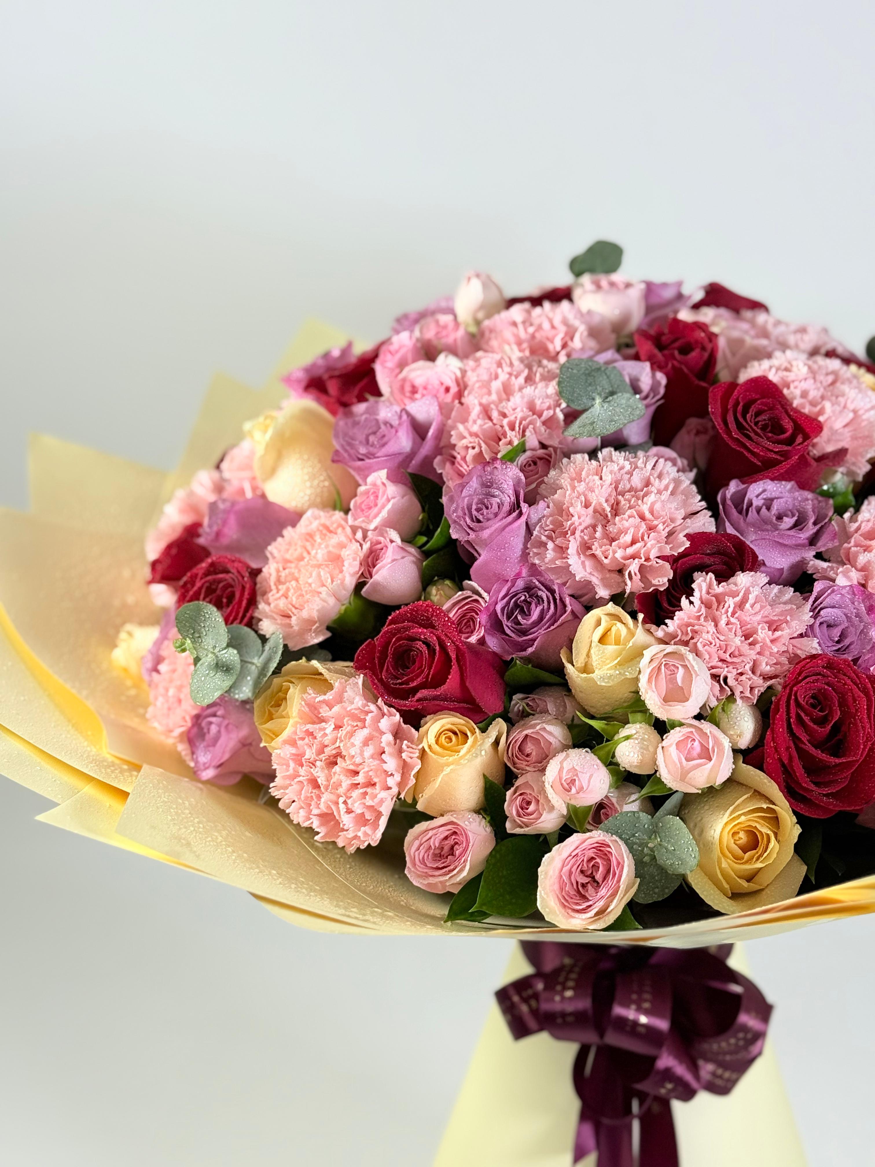 Vibrant bouquet of pink, red, and yellow roses with carnations, wrapped in yellow paper and purple ribbon.