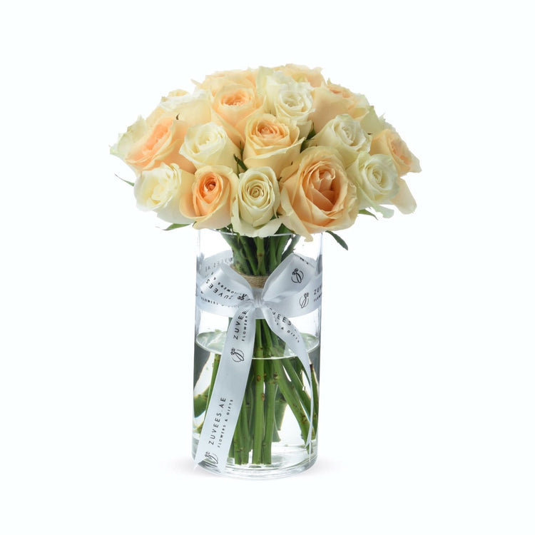 Soft peach and cream roses in clear glass vase with branded white ribbon.