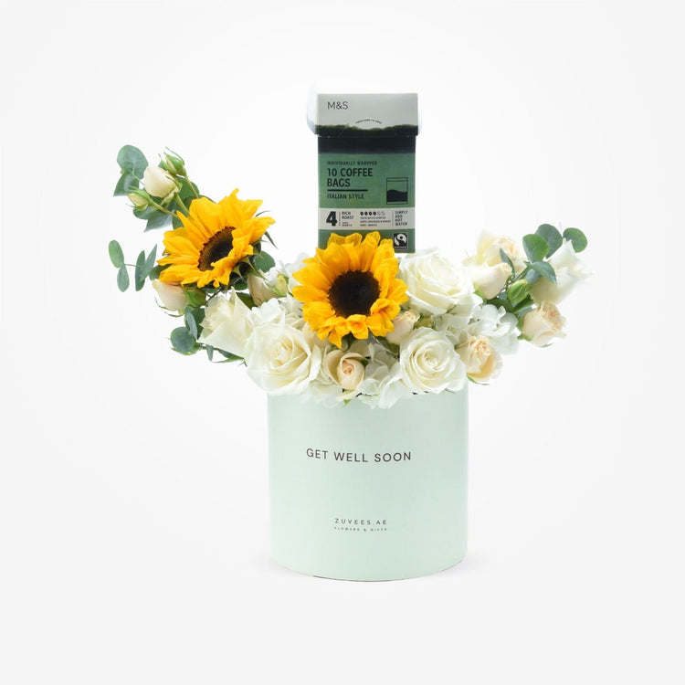 Pastel mint cylinder holds white roses and sunflowers, topped with M&S coffee, "Get Well Soon" message.