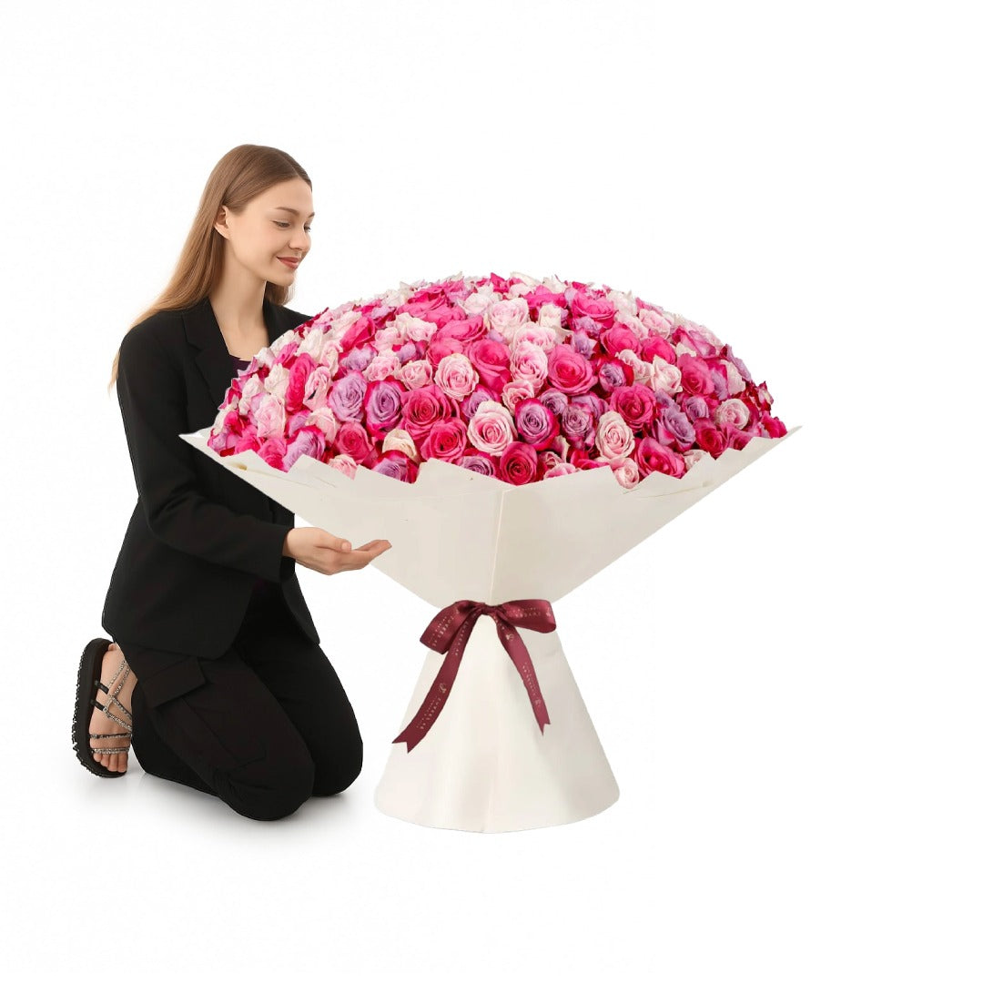 Woman kneels beside massive, vibrant pink rose bouquet in elegant white wrap with burgundy bow.