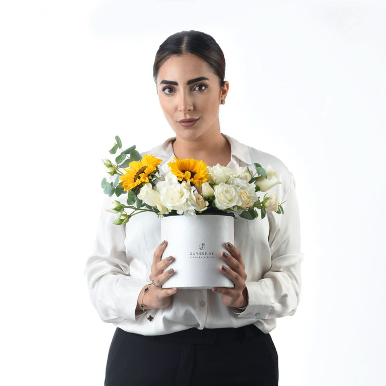 Woman holds elegant white hat box filled with sunflowers and roses against clean white background.