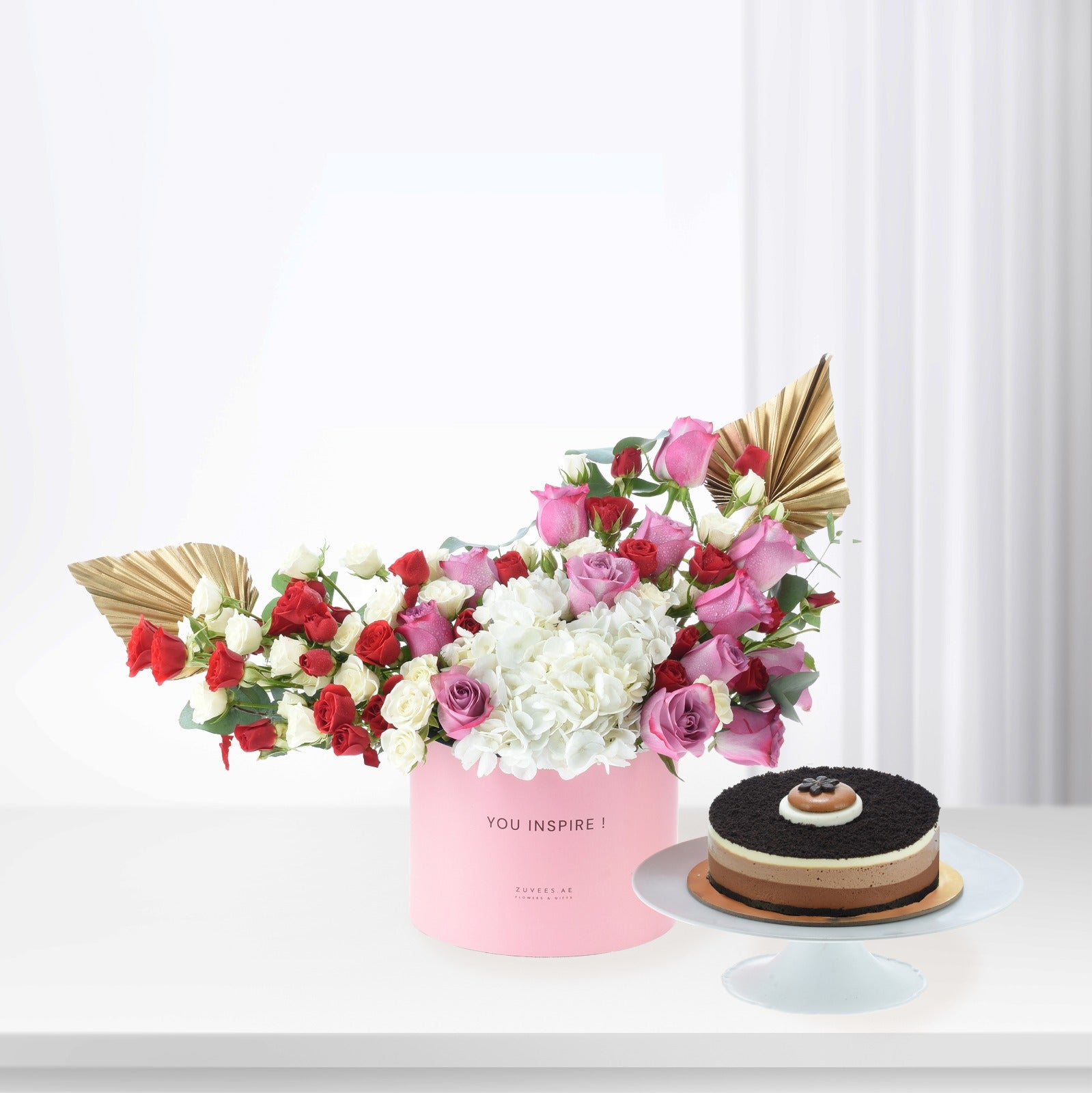 Pink box holds vibrant red, pink, white roses and hydrangeas, accented with gold fans. Beside, layered chocolate cake on white stand.