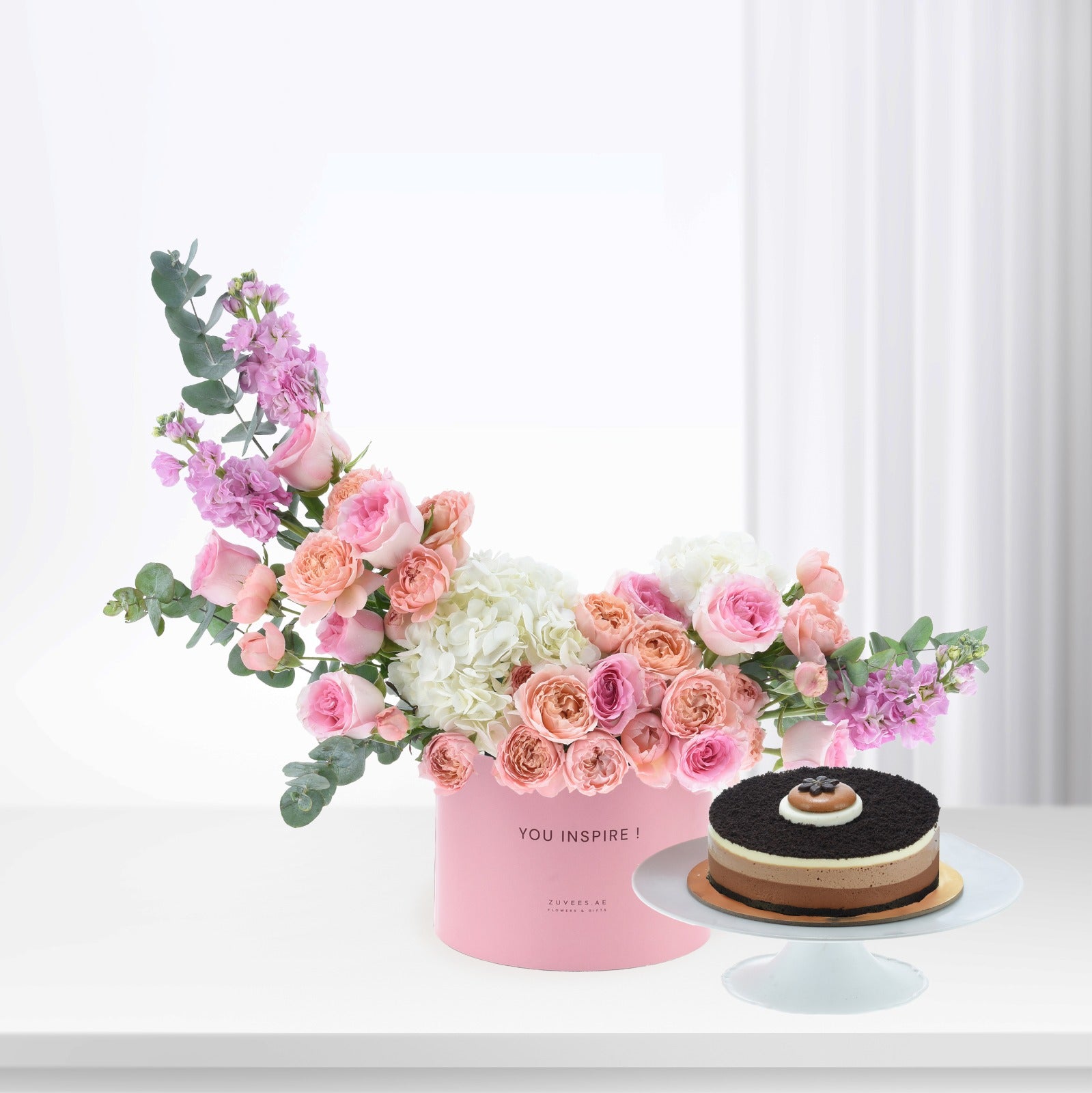 Pink floral arrangement with roses, hydrangeas, eucalyptus in "YOU INSPIRE!" box beside chocolate cake.