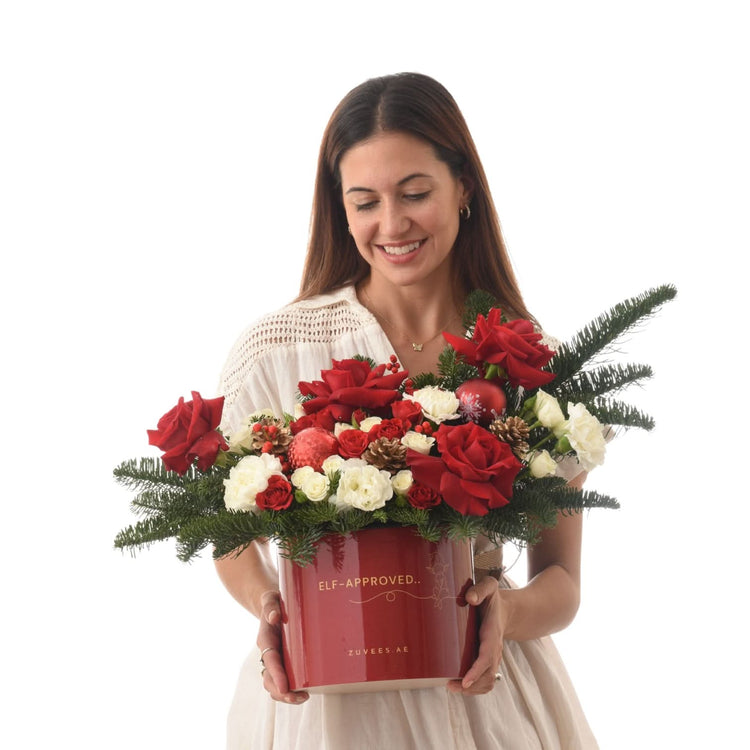 Woman holds festive red-and-white floral arrangement in glossy "Elf-Approved" box.