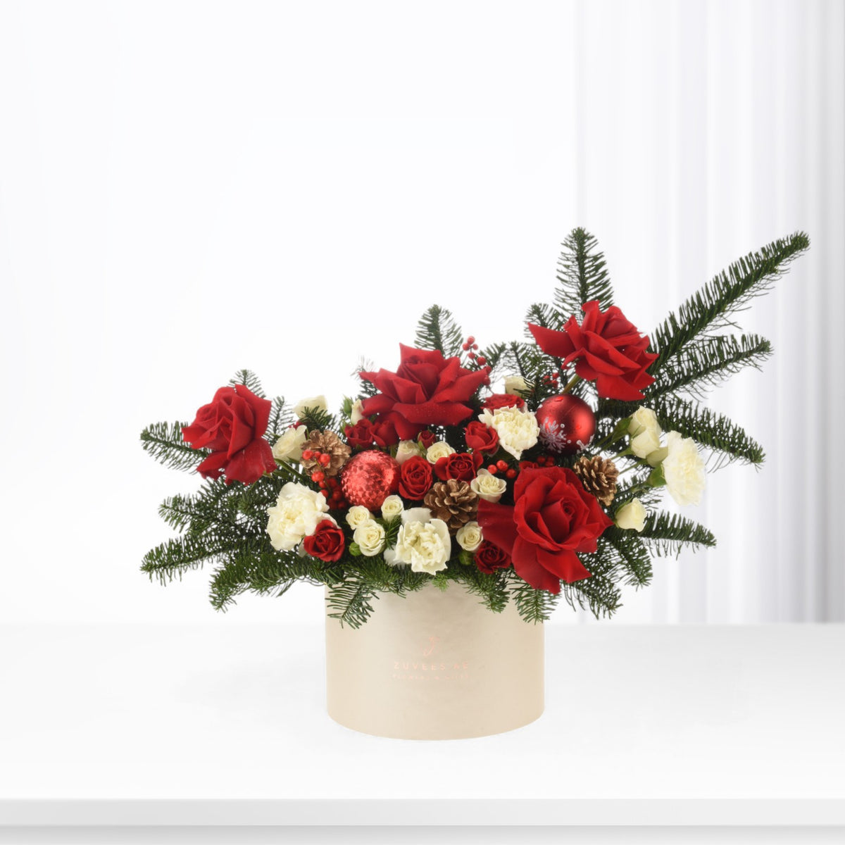 Vibrant red roses, white carnations, pine, and ornaments in elegant beige box.