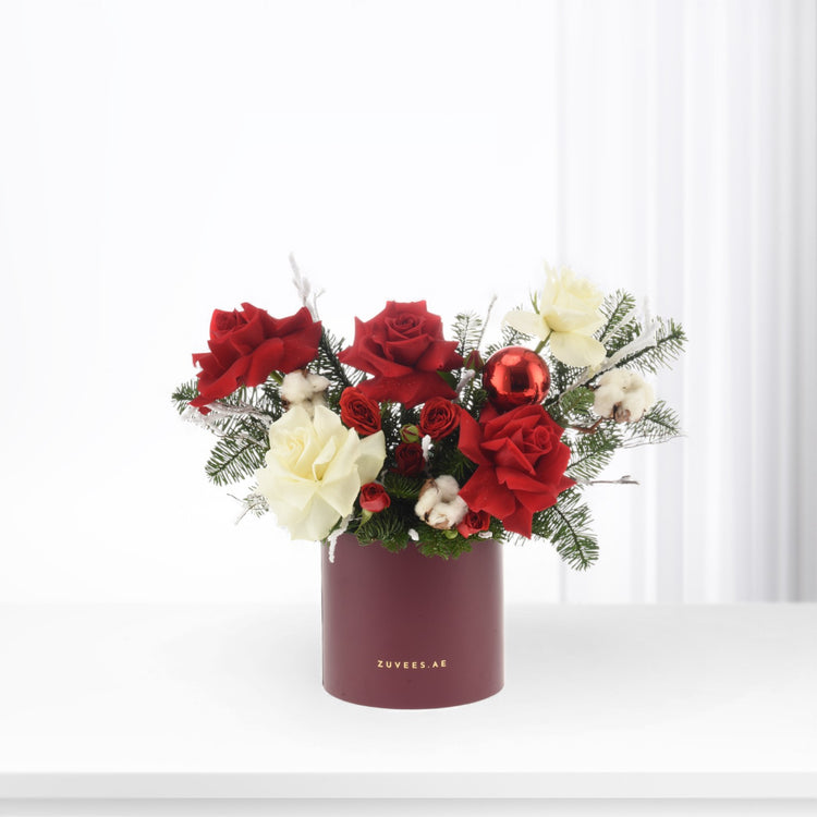 Vibrant red and white roses, cotton, pine, and a glossy red ornament in a burgundy cylinder vase.
